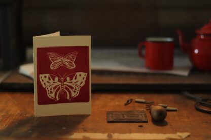 Handbedruckte Karte mit einem Linoldruck, der zwei Schmetterlinge zeigt, Im Hintergrund Werkstatt und Druckerpresse