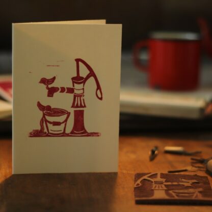 Handbedruckte Karte mit einem Linolschnitt von einer Schwengelpumpe mit Eimer und badenden Vögelchen, Werkstattsituation mit Linolplatte und Schnittwerkzeug, im Hintergrund die Druckerpresse
