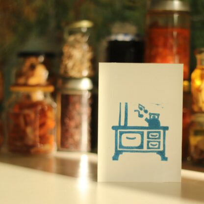 Handbedruckte Karte mit einem Linolschnitt, der eine alte Küchenhexe zeigt auf der ein Wasserkessel singt, im Hintergrund Gläser mit Eingemachtem