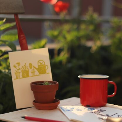 Arbeitssituation: Linolschnittwerkzueg, Stifte, fertige handbedruckte Kartenb mit Linolschnitten auf Gartenstuhl, im Hintergrunf Pflanzen