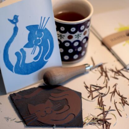 Werkstattsituation: Linolplatte und Linolschnittwerkzeug, sowie fertig gedrucktes Motiv das ein Kätzchen mit einem Vögelchen zeigt