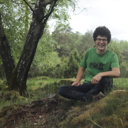 Junger Mann sietzt auf grüner Wiese, mit grünen T-shirt auf dem drei Kühe gedruckt sind, im Hintergrund Natur