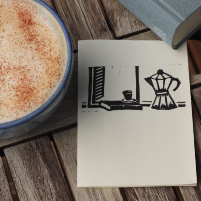 Handbedruckte Karte mit einem Linolschnitt der eine Espressokanne und ein Buch mit Tässchen im offenen Fenster zeigt, daneben ein Kaffe mit Milchschaum und ein Buch