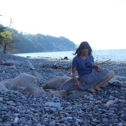 Frau an Steinstrand mit Rock und blauem T-Shirt, auf dem der Linolschnitt einer dicken Meerjungfrau gedruckt ist