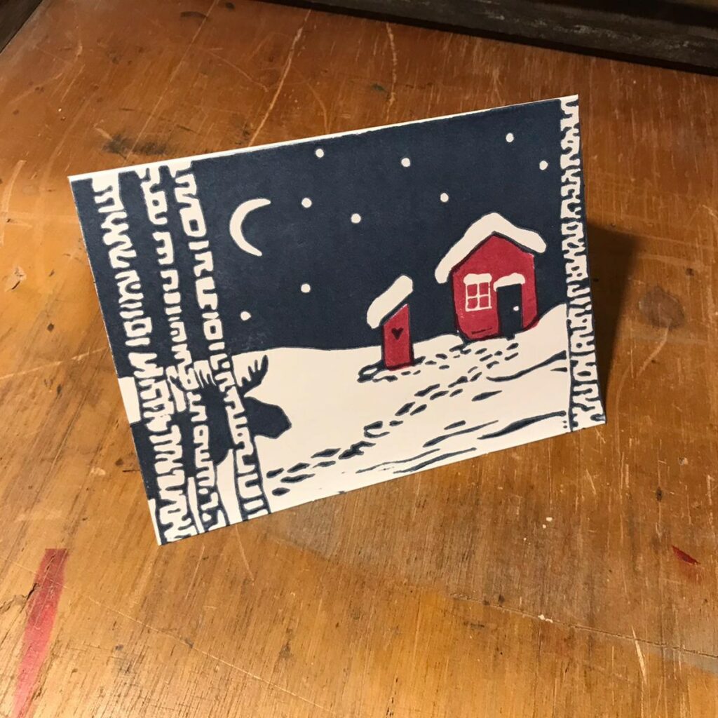 Handbedruckte Weihnachtskarte mit einem Linoldruck, der ein rotes Schedenhaus im Schnee zeigt, daneben ein rotes Plumpsklo mit Fußstapfen im Schnee, im Vordergrund Birken und ein Elch